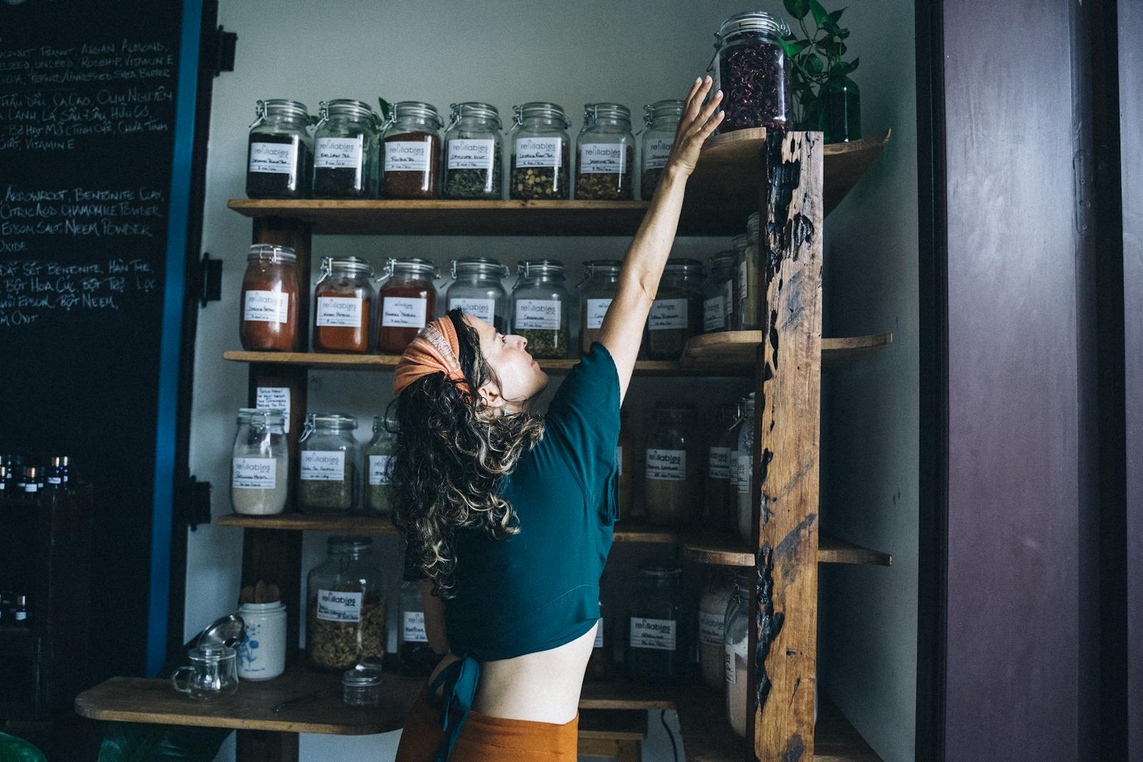 Woman reaching for a jar on a shelf in an eco-friendly shop with wooden decor and glass containers.