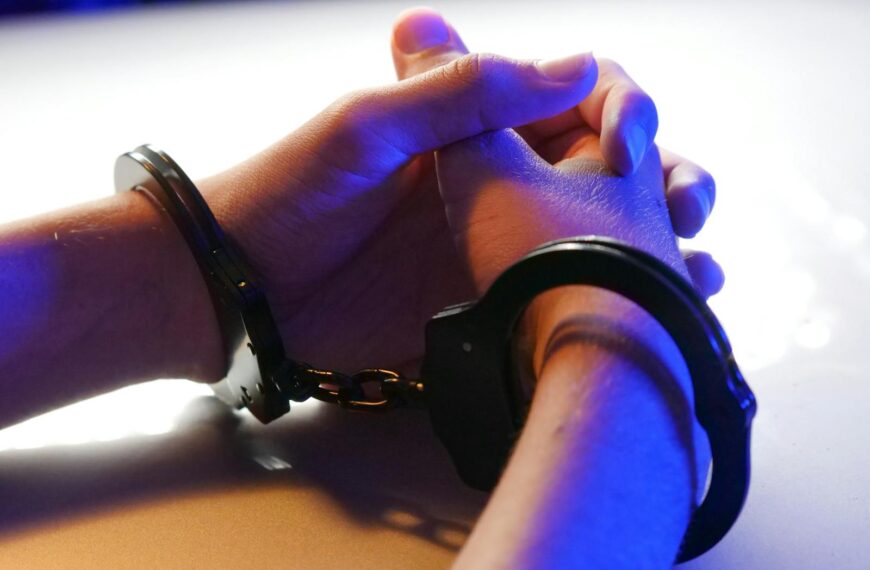 Close-up of handcuffed hands under blue police light, signifying arrest or detention.