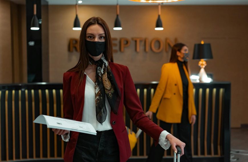 A woman in a mask stands at a hotel reception desk holding paper, welcoming service