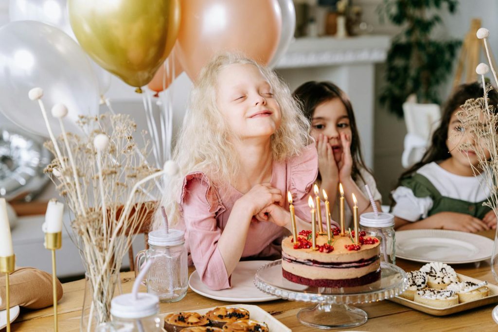 A happy girl makes a wish with lighted candles on her birthday cake, surrounded by friends and balloons.