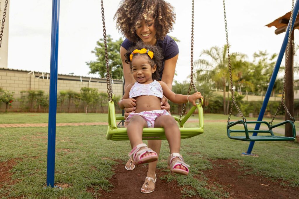 A happy mom pushes her daughter on a swing in a sunny playground, enjoying a beautiful day.