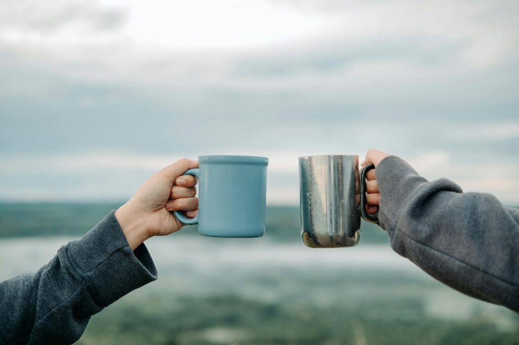 Two hands holding mugs outdoors with a scenic blurry background, symbolizing friendship and relaxation.