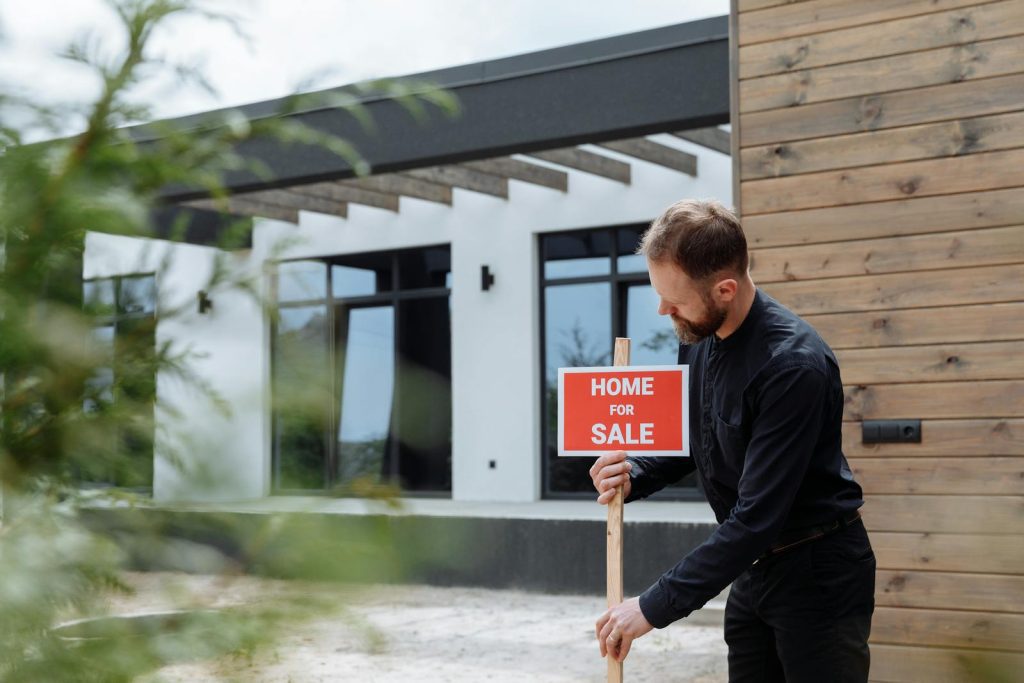 Man placing a home for sale sign outside a modern house for real estate listing.