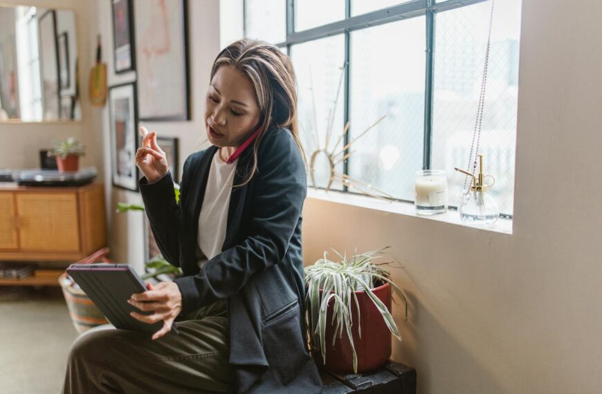 Asian woman multitasks at home using a tablet and phone in a cozy setting.
