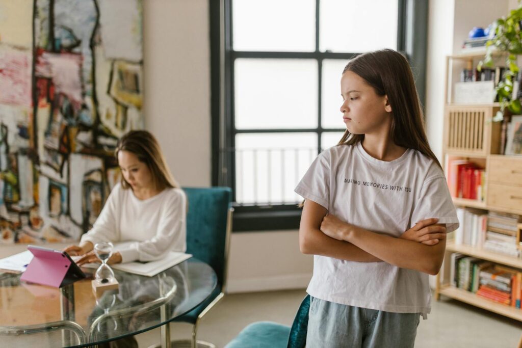 A young girl crosses her arms, appearing upset, while her mother works at home.