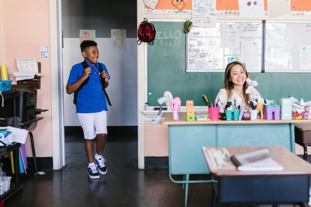 A cheerful student enters the classroom where a teacher is seated at a desk, ready for class.