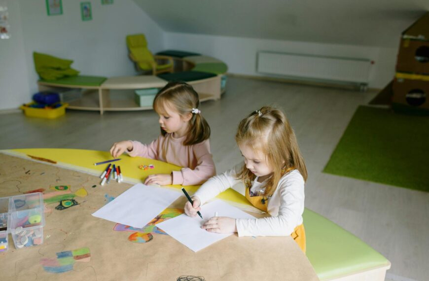 Two young girls in a classroom, absorbed in drawing with colorful markers and paper.