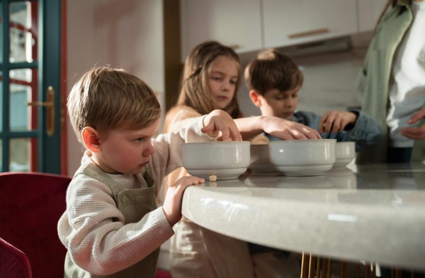 Three children gather around a table enjoying breakfast indoors on a sunny morning.