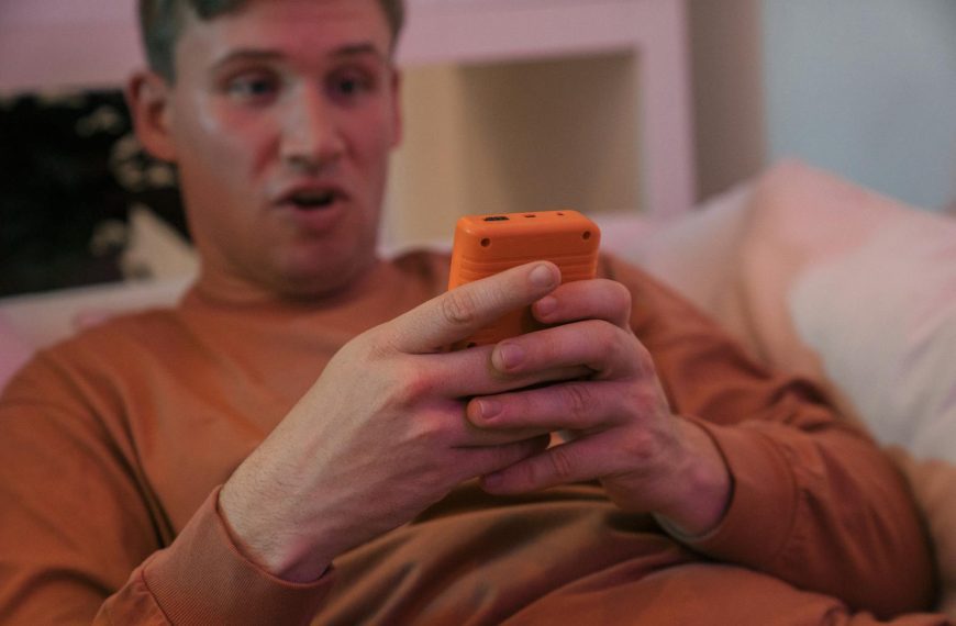 A man lying in bed looking surprised while using a smartphone indoors.