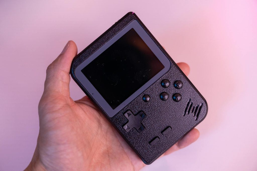 Close-up of a hand holding a black retro handheld game console against a pink background.