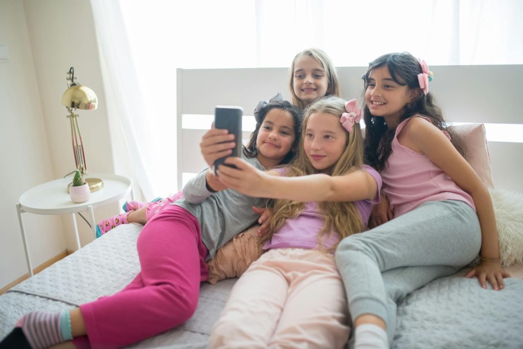 Four young girls in pajamas enjoy a joyful sleepover, capturing moments with a smartphone selfie.
