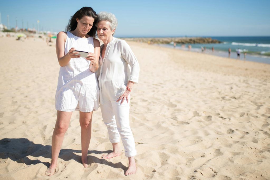 Mother and daughter enjoying a sunny day at a Portuguese beach, sharing moments on a smartphone.