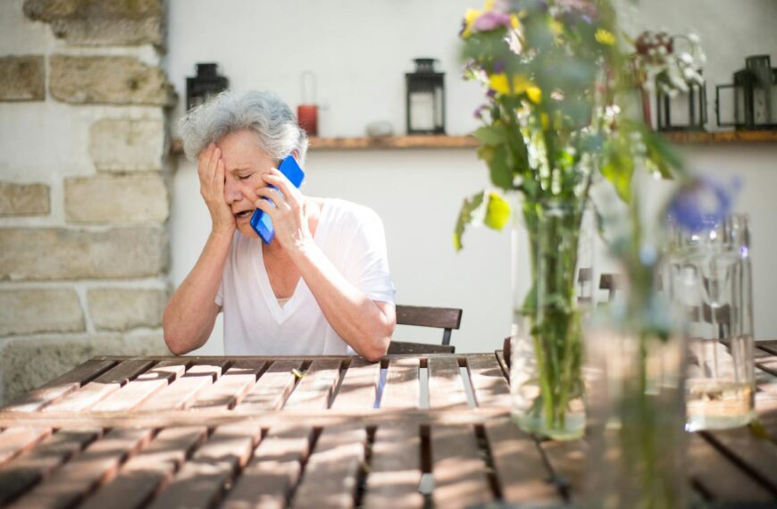 A distressed senior woman sitting outdoors, holding a cellphone, and showing emotion.
