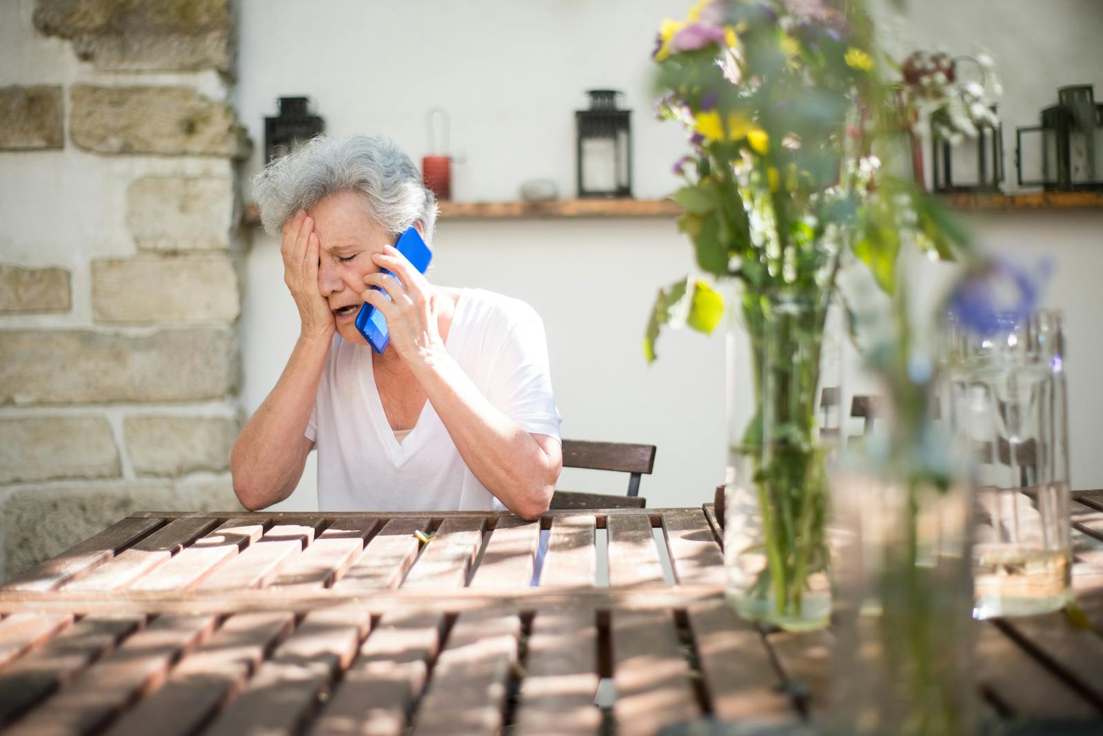 A distressed senior woman sitting outdoors, holding a cellphone, and showing emotion.