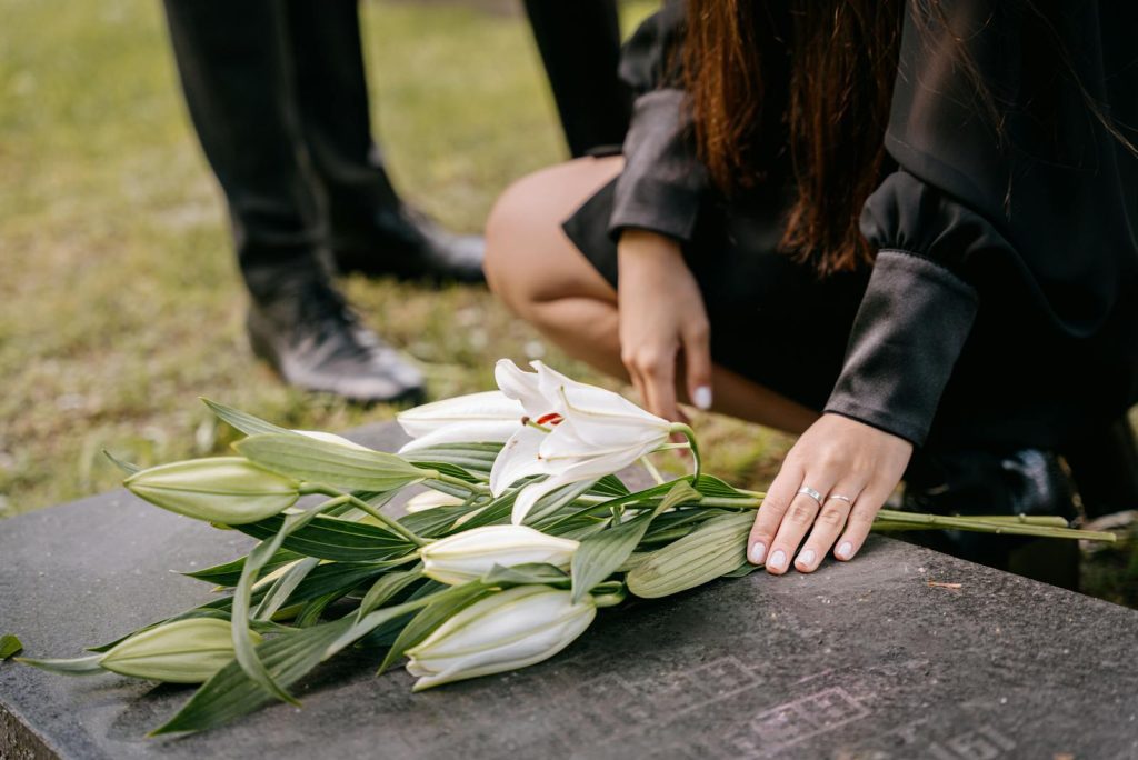 A woman in black attire mourns beside a grave, placing white lilies on the headstone in a cemetery setting.