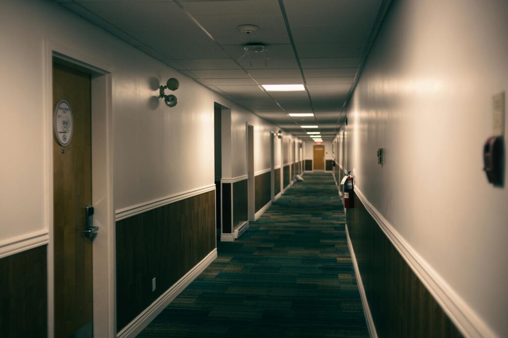 A quiet hotel hallway with carpeted flooring and wooden doors, illuminated by ceiling lights.