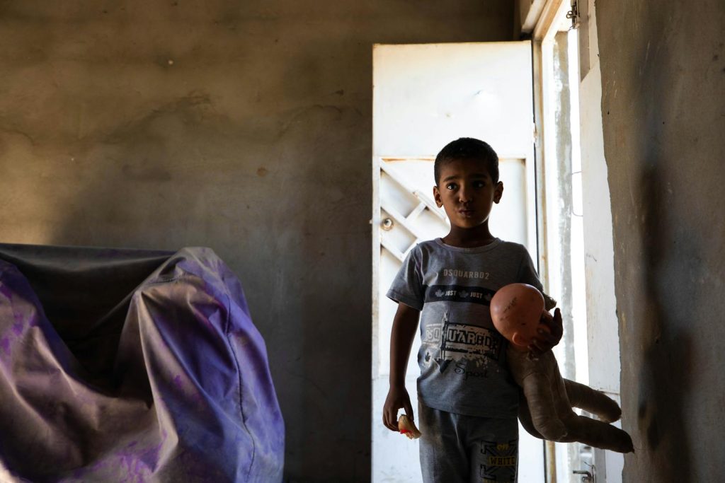 A child standing in a shadowy room, holding a toy doll with a somber expression.