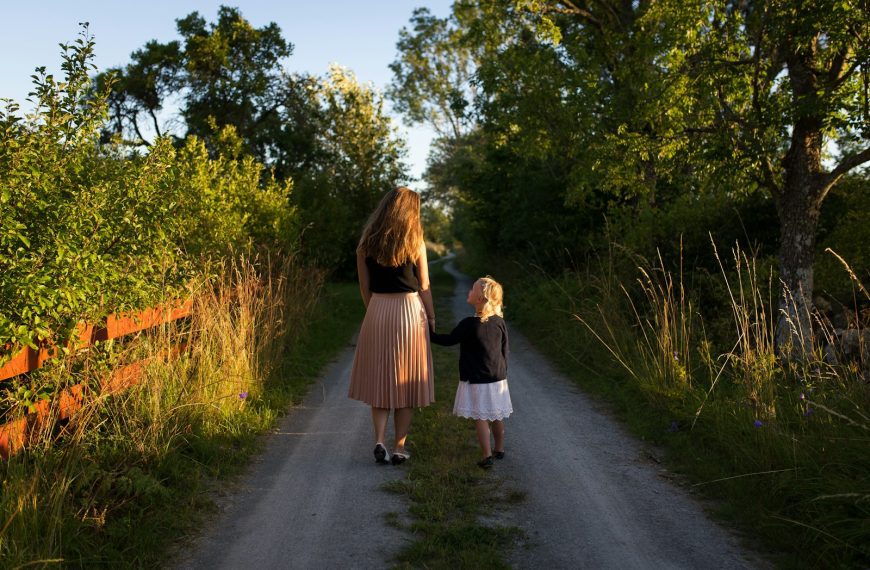 woman and girl walking on road surrounded by green grass