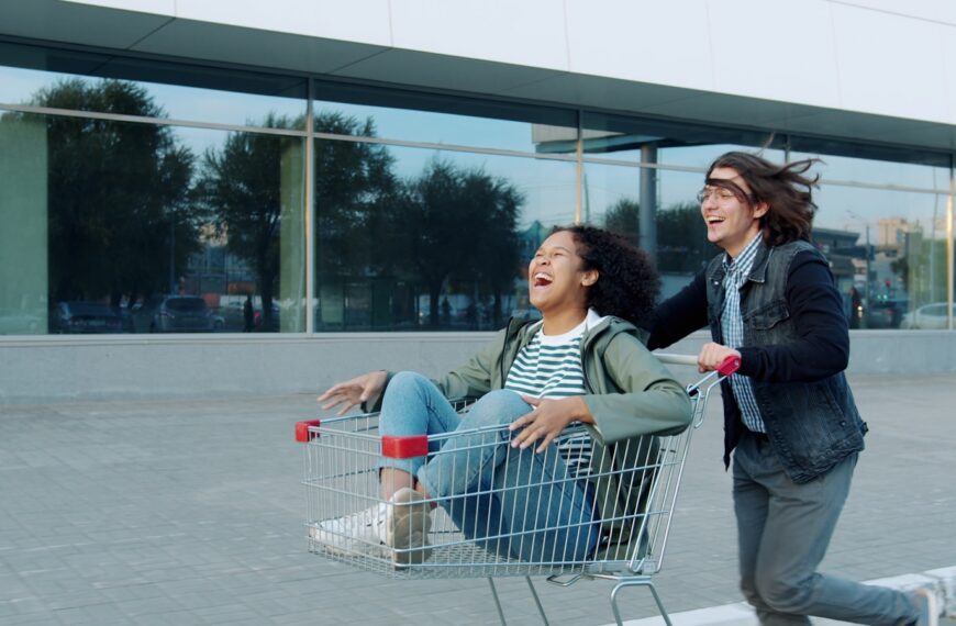 Two people riding in a shopping cart outside.