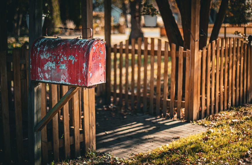 A weathered red mailbox stands by a wooden fence.