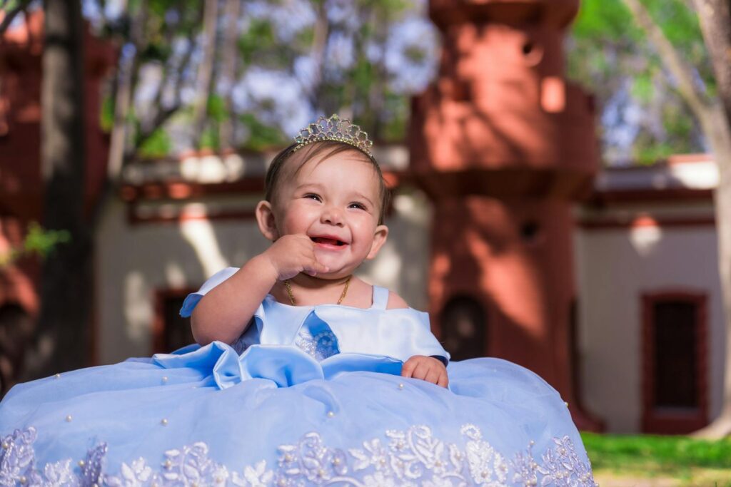 Charming baby girl dressed as a princess with a tiara, smiling joyfully outside.