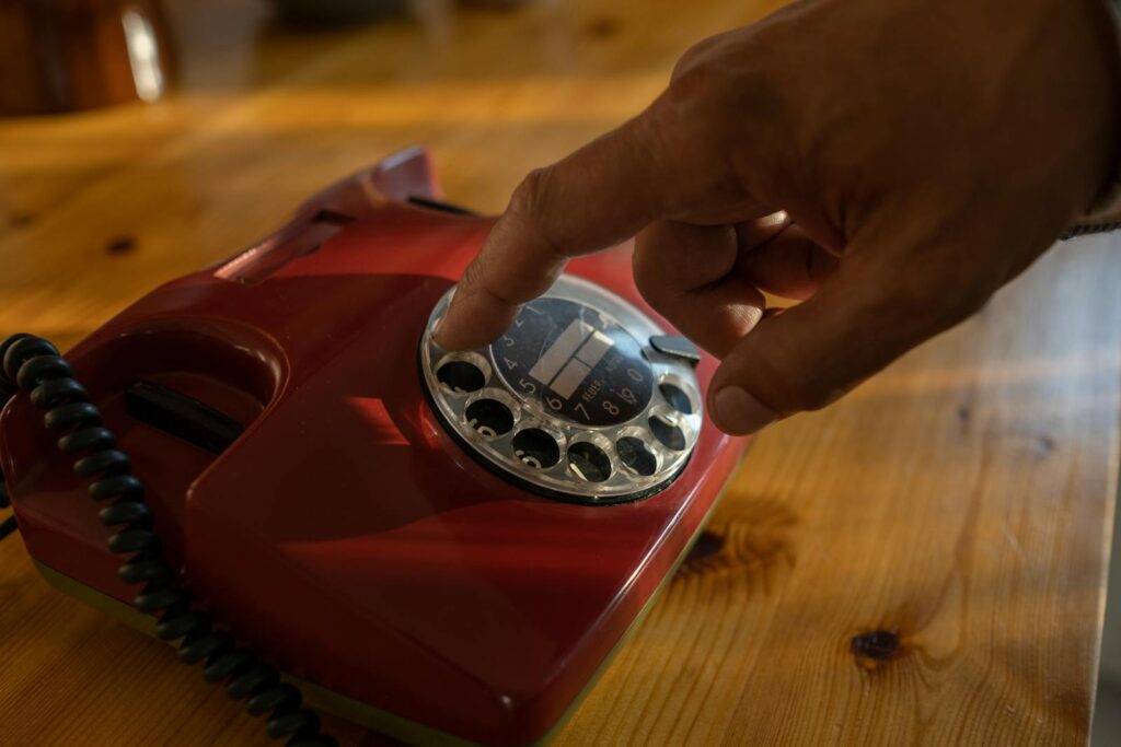 Close-up of a hand dialing a red rotary phone on a wooden table.