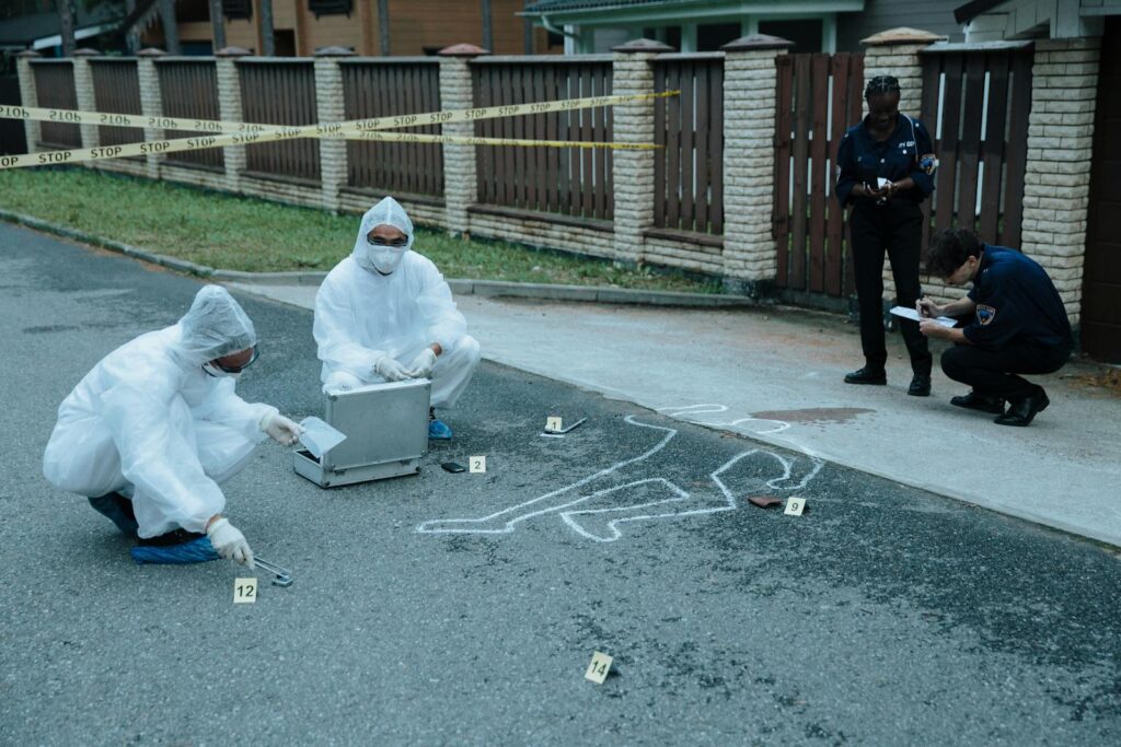 Police officers and investigators examine a crime scene with evidence markers and chalk outline.