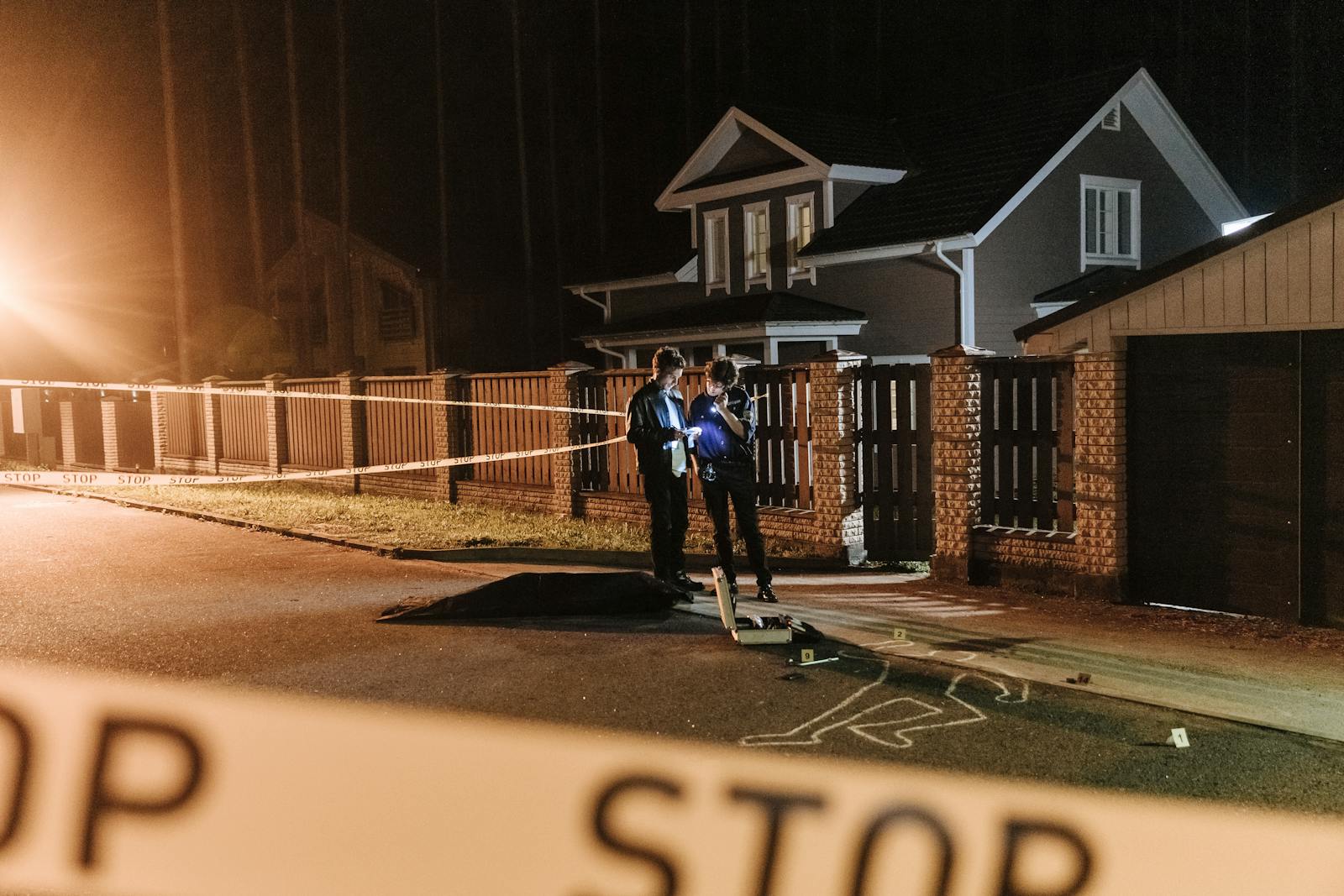 Police officers investigating a nighttime crime scene outside a residential house with barrier tape and evidence markers.