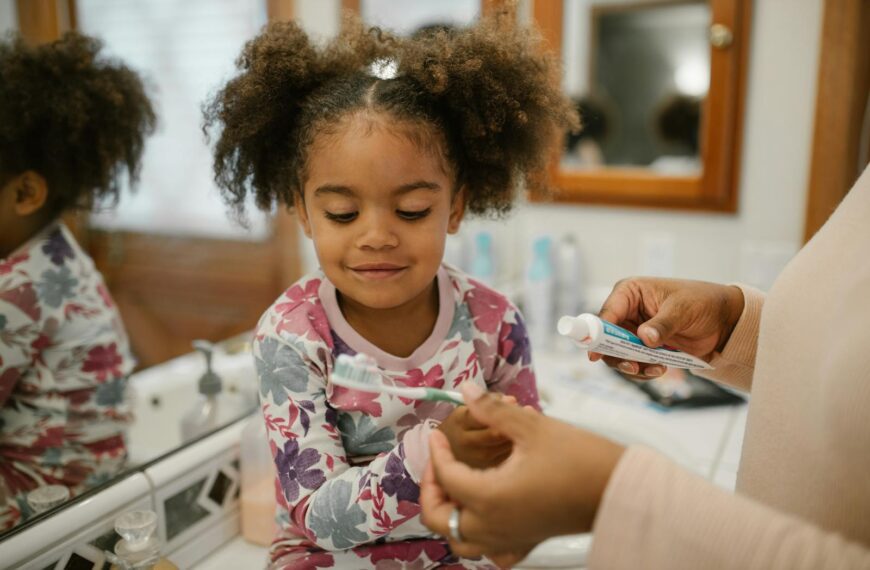 A mother assists her daughter in brushing her teeth in a cozy, home bathroom setting.