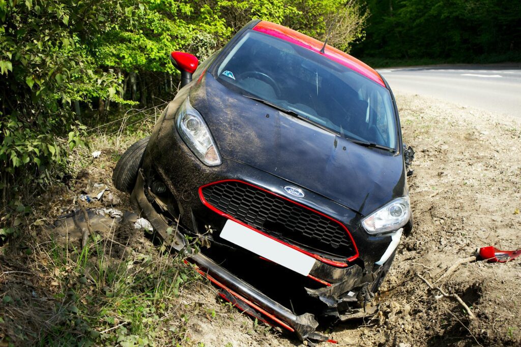 A black car, seemingly a Ford, crashed and damaged in a roadside ditch with surrounding greenery.