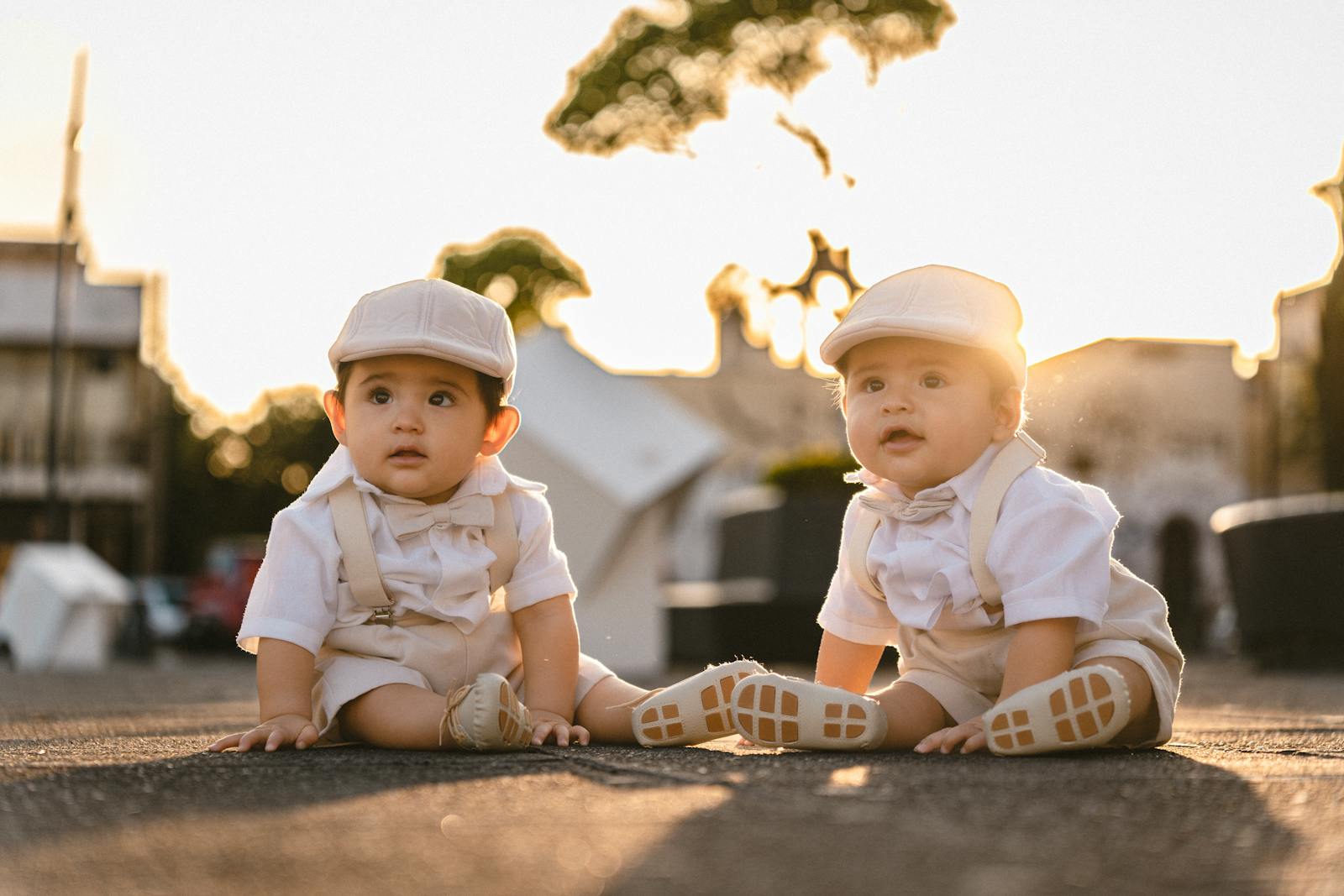 Adorable twin babies dressed in elegant attire sitting outdoors at sunset, creating a warm and cute scene.