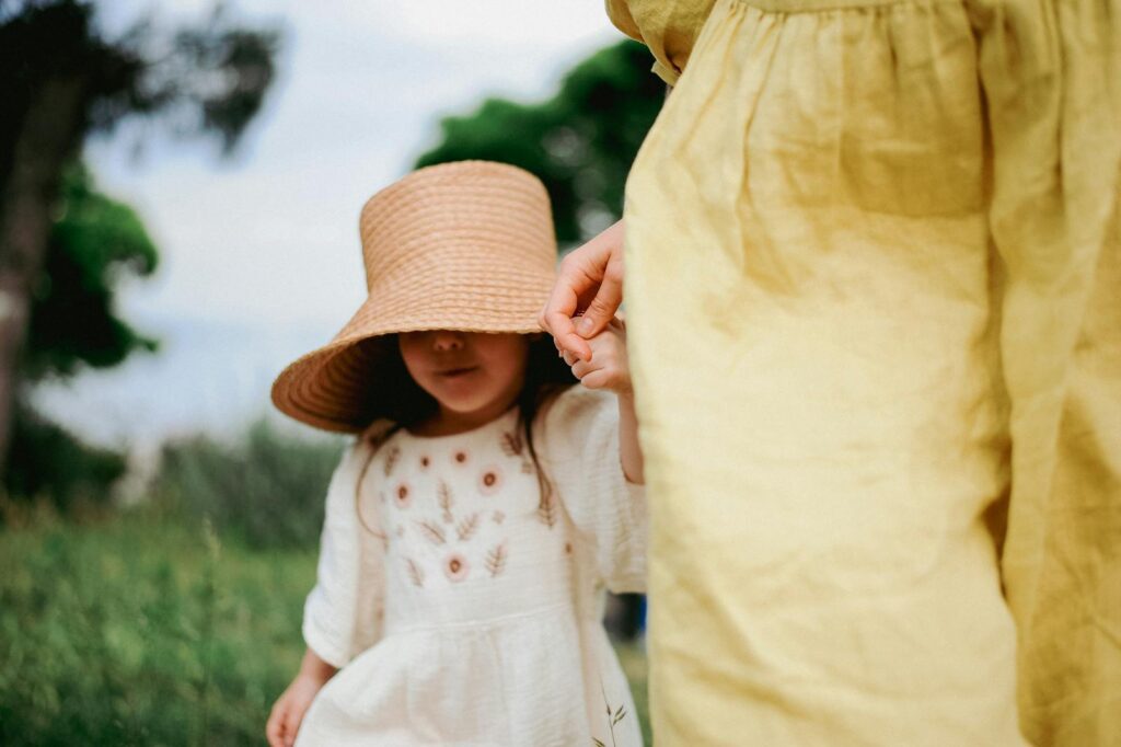 A little girl in a white dress and straw hat holds her mother's hand outdoors.