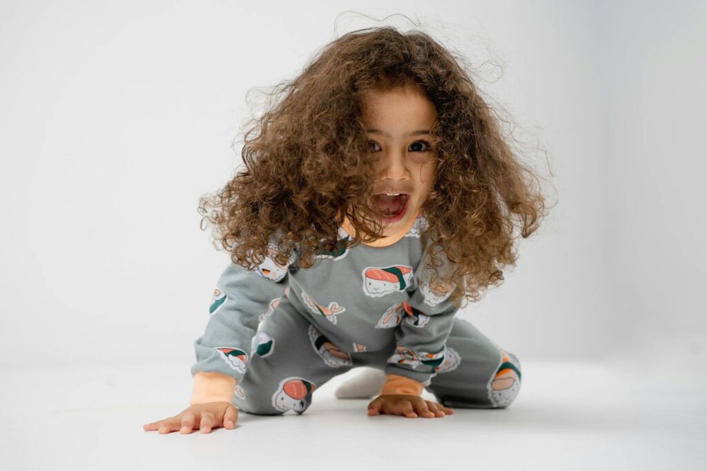 A happy child wearing playful pajamas with long brown hair, kneeling and smiling on a white background.