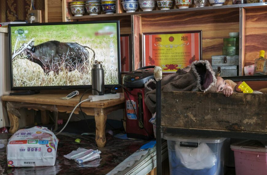 A baby in a wooden cot next to a TV in a warm, homely interior setting.