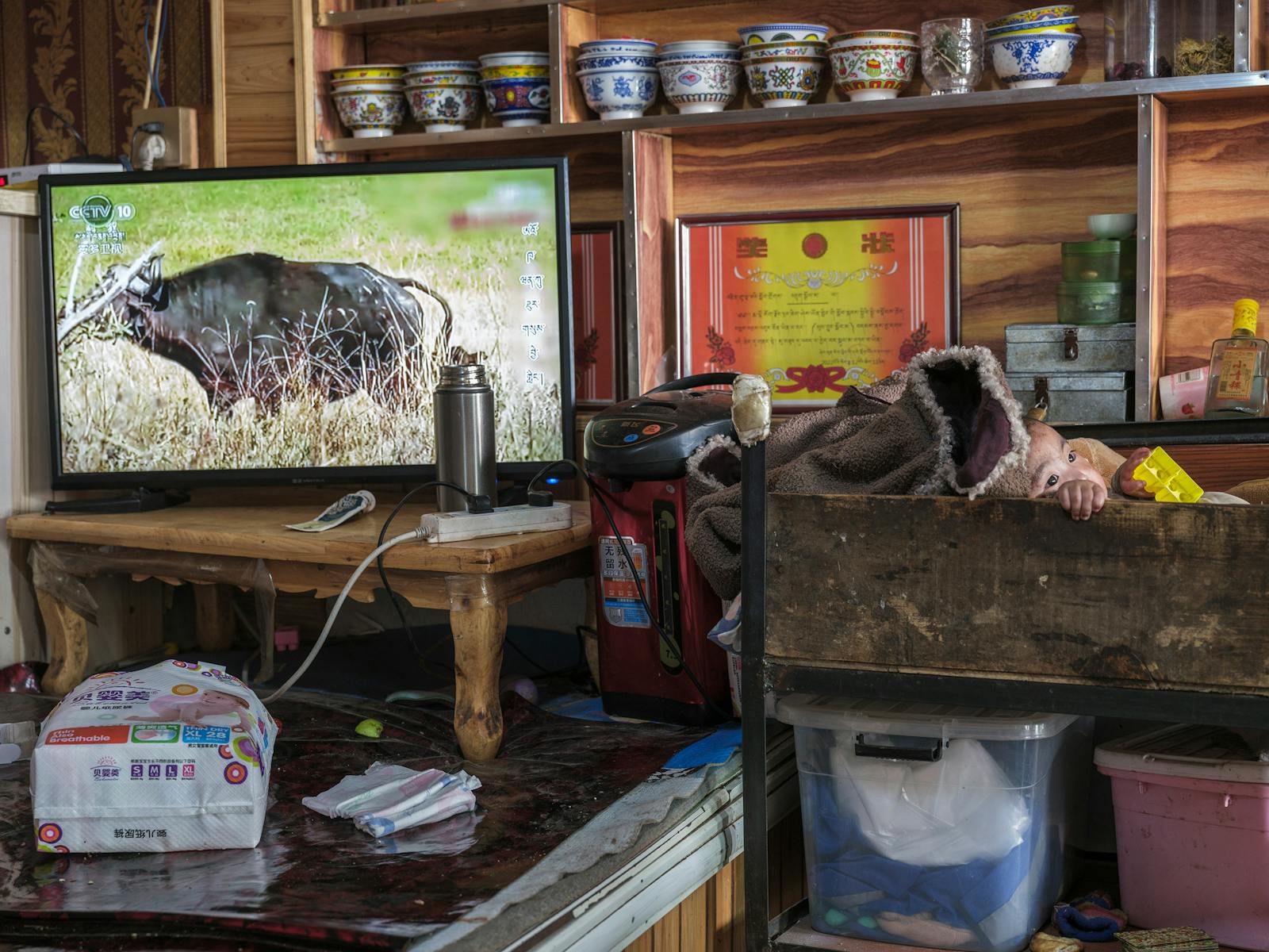 A baby in a wooden cot next to a TV in a warm, homely interior setting.