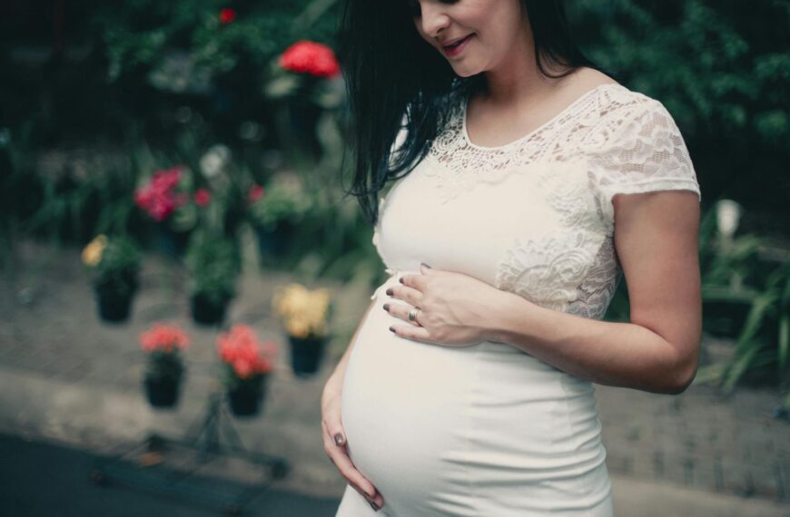Pregnant woman in white lace dress, posing outdoors with flowers, exuding happiness.