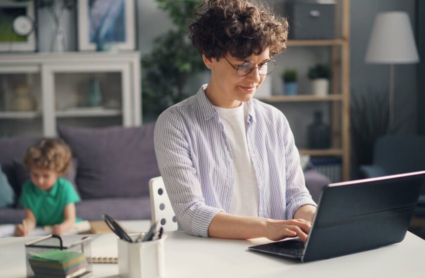 A mother working remotely from home on her laptop while her child plays in the background.