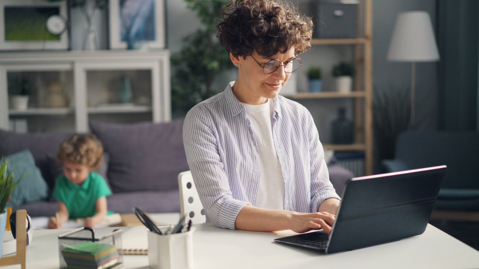 A mother working remotely from home on her laptop while her child plays in the background.