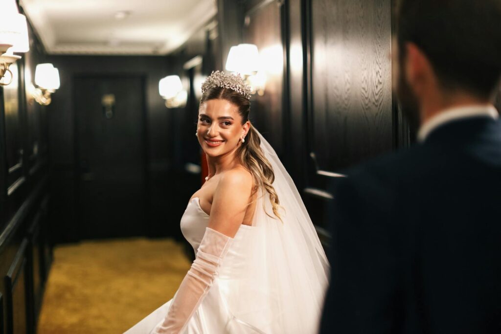 Bride in dress and tiara smiling at groom in elegant hotel hallway, capturing a joyful wedding moment.