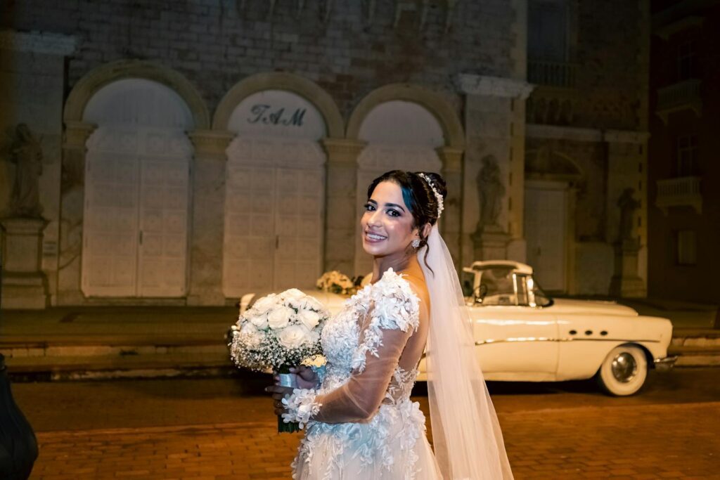 Bride in lace dress posing with classic car at night, smiling and holding bouquet.