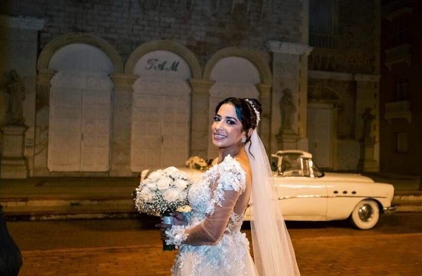 Bride in lace dress posing with classic car at night, smiling and holding bouquet.
