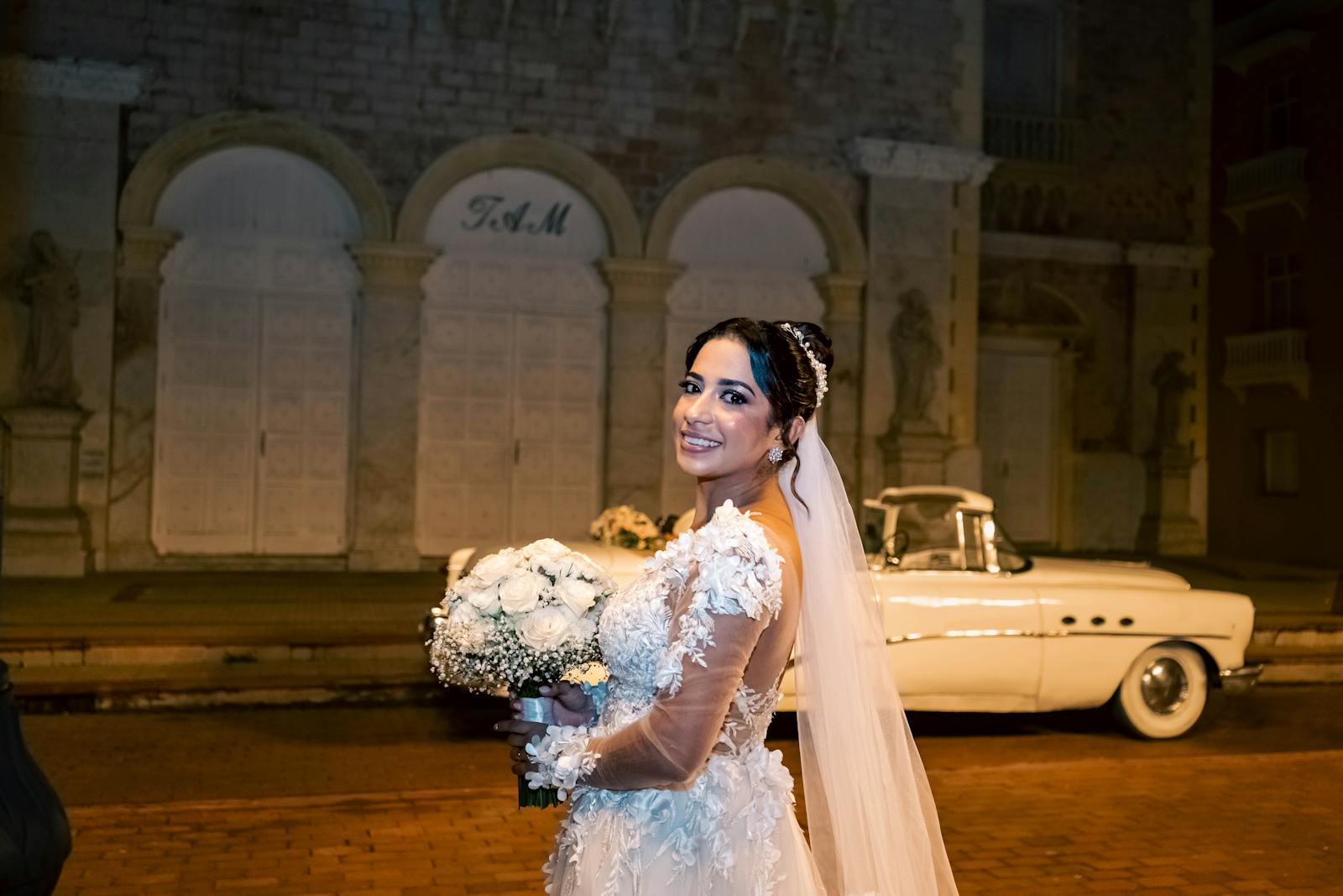 Bride in lace dress posing with classic car at night, smiling and holding bouquet.
