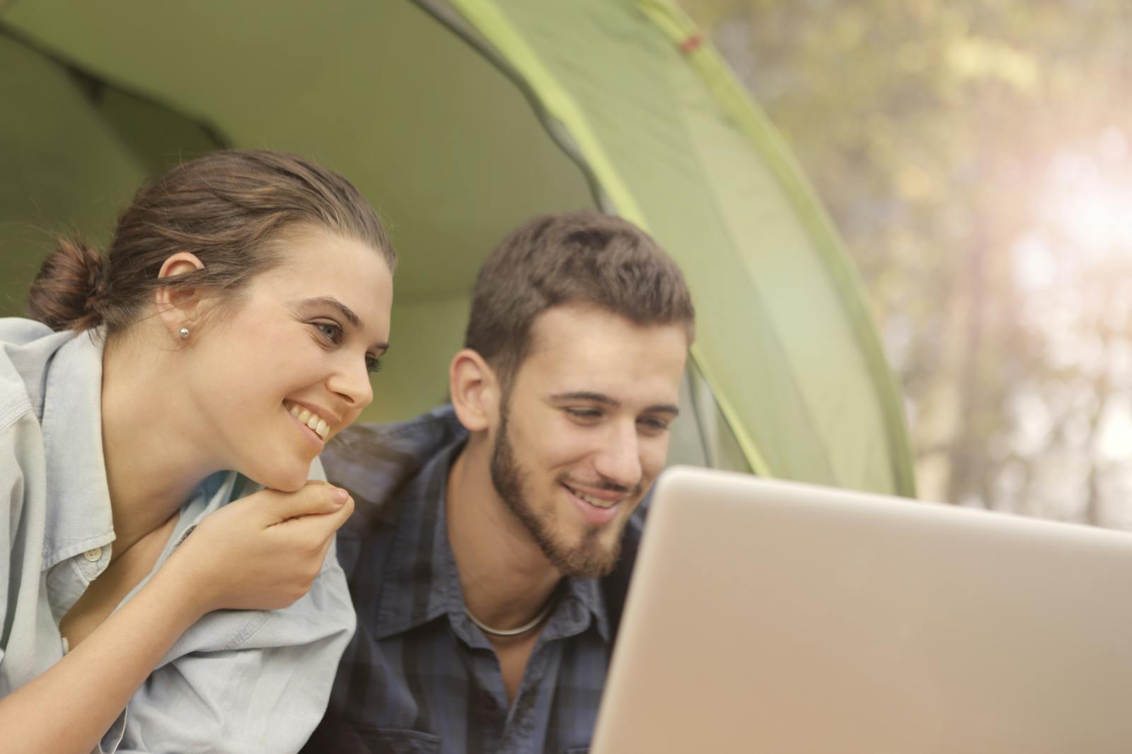 A happy couple in a tent enjoying leisure time with a laptop in a sunny outdoor setting.