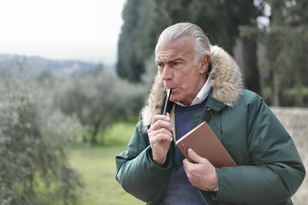 A senior man with grey hair contemplates thoughtfully while holding a book and pen outdoors.