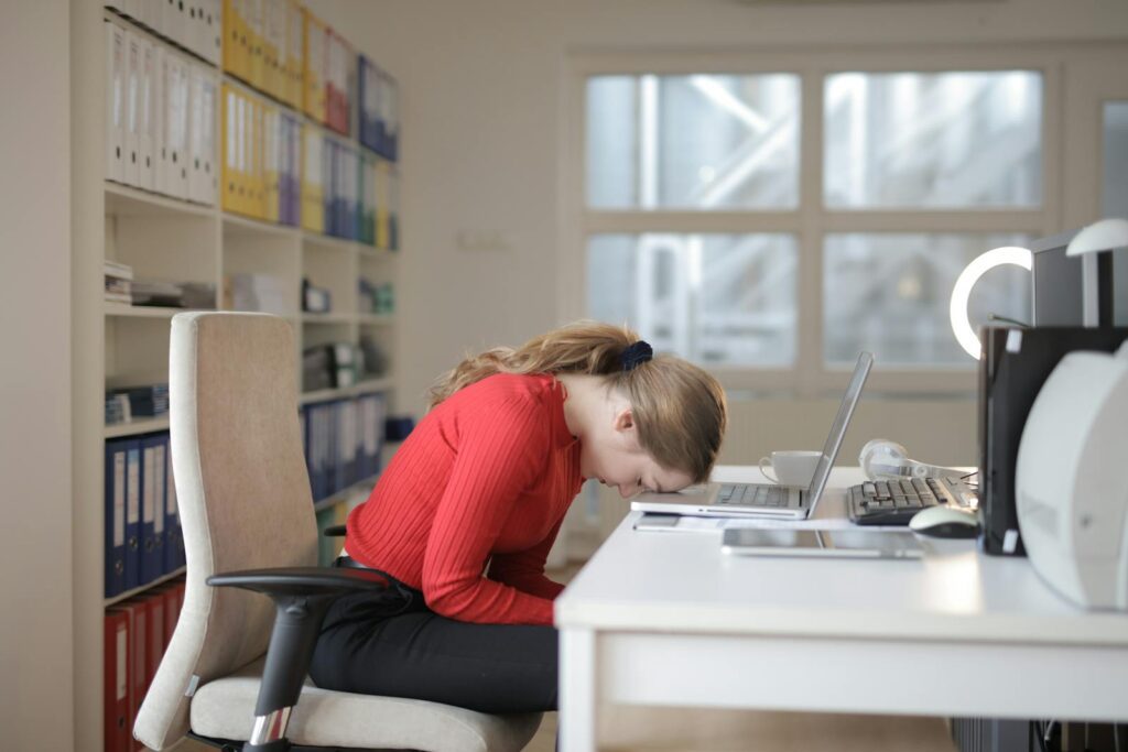 Tired woman in red sweater naps on office desk beside laptop, overwhelmed by remote work.