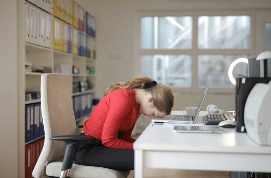 Tired woman in red sweater naps on office desk beside laptop, overwhelmed by remote work.