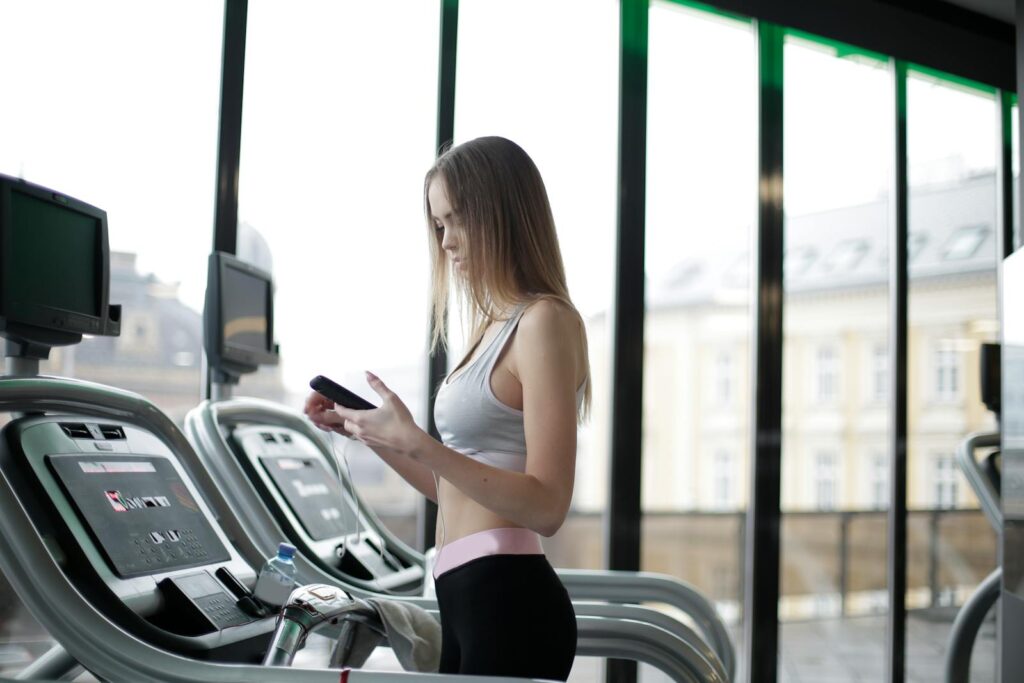 Side view of slim young female athlete in activewear using smartphone while training on treadmill in spacious modern fitness club
