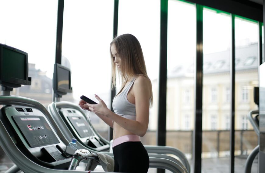 Side view of slim young female athlete in activewear using smartphone while training on treadmill in spacious modern fitness club