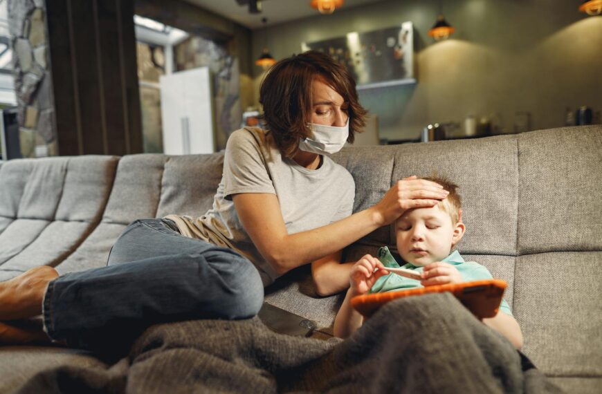 A concerned mother wearing a mask checks her child's temperature on the sofa.