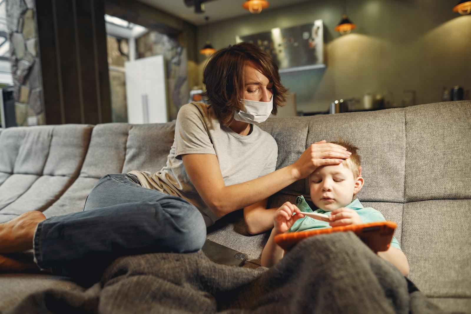 A concerned mother wearing a mask checks her child's temperature on the sofa.
